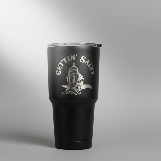 A matte black insulated travel tumbler with a clear lid, featuring a silver engraved graphic and the words 'GETTIN' SALTY' on the front, displayed on a white rectangular box against a gray studio background. alternate view