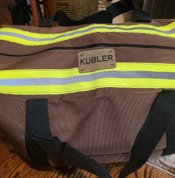 Brown bunker gear bag with reflective stripes and a personalized name label on a wooden surface.