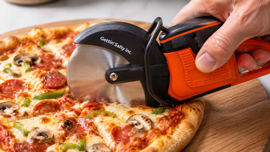 A close-up of a hand using an orange and black Gettin' Salty pizza cutter to slice a pepperoni and mushroom pizza on a wooden board.