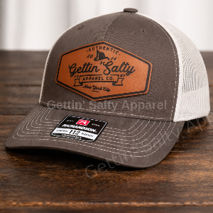 A brown and white trucker hat with a brown leather patch on the front that reads 'Gettin' Salty Apparel Co.,' displayed on a dark wooden surface.
