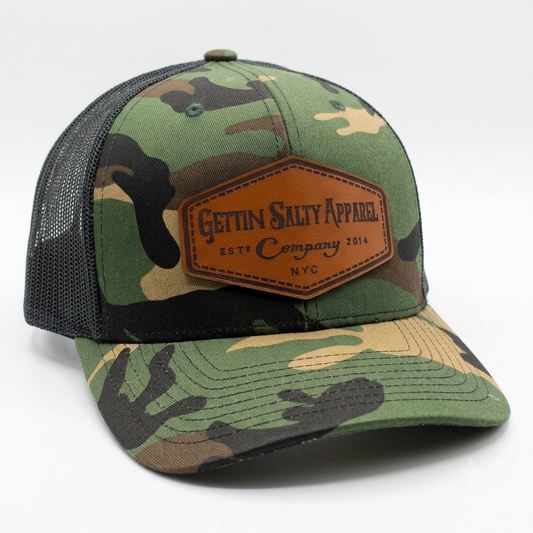 A camouflage trucker hat with a brown leather patch on the front that reads 'Gettin' Salty Apparel'. The hat features green, brown, and black camo on the front panel and visor, with a black mesh back.
