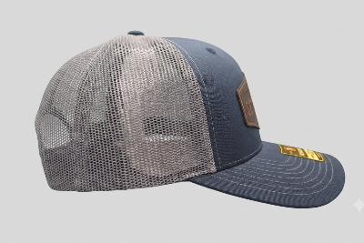 A trucker-style baseball cap with a navy blue front panel and brim, gray mesh back panels, a brown leather patch on the side, and a gold sticker on the brim, shown against a light gray background. alternate view