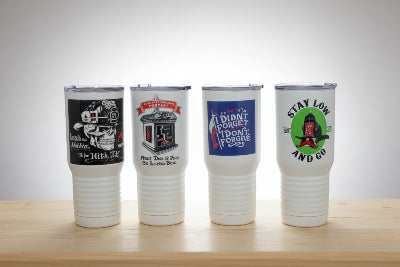 Four white insulated travel tumbler cups with printed graphics, lined up on a light wood surface against a neutral gray background.
